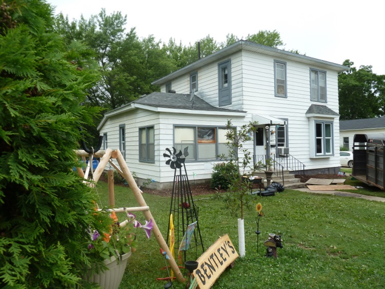 White two-story house with a porch and garden decorations, including a sign reading 