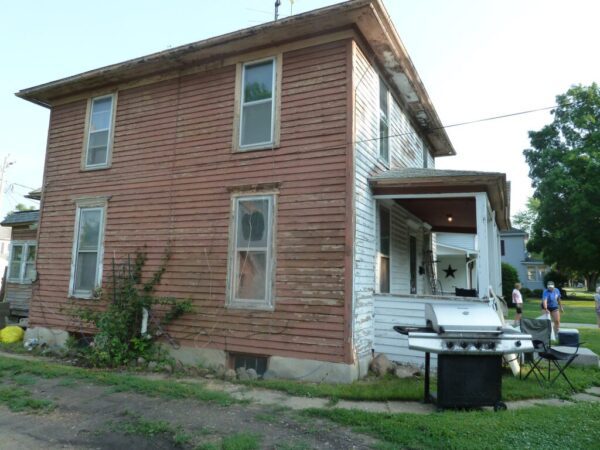 Two-story weathered wooden house with faded siding and a porch. A gas grill stands nearby. People and trees are visible in the background.