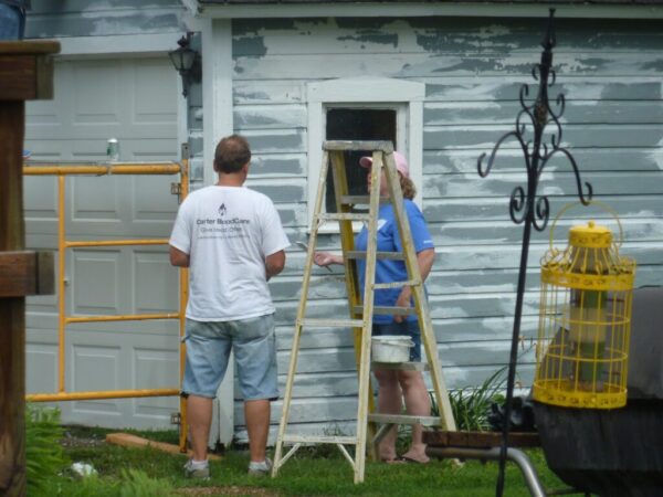 Two people painting an old wooden building's exterior. One stands on a ladder, wearing a cap. The other, in a white t-shirt, stands nearby. Scaffolding and garden objects are visible.