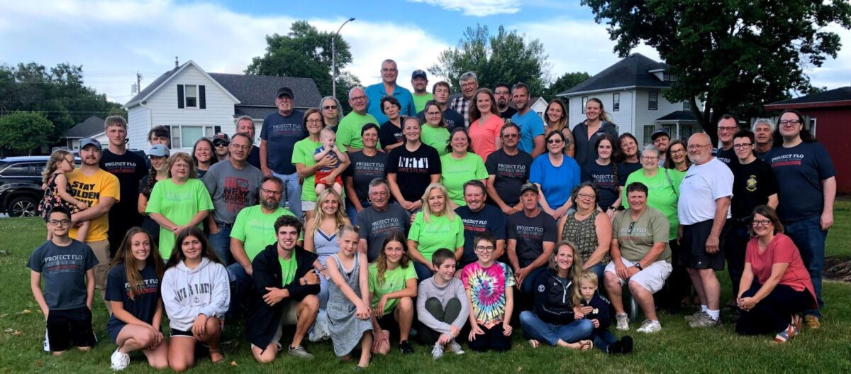 A large group of people, including adults and children, are gathered outdoors on grass, some wearing matching shirts. Trees and houses are in the background.