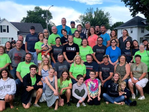 A large group of people, including adults and children, are gathered outdoors on grass, some wearing matching shirts. Trees and houses are in the background.