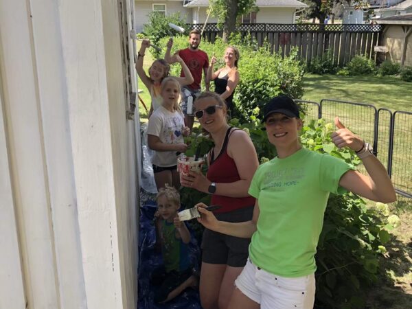 A group of people, including adults and children, paint a white wooden wall outdoors in a garden setting. One person gives a thumbs-up, and tarps cover the ground.