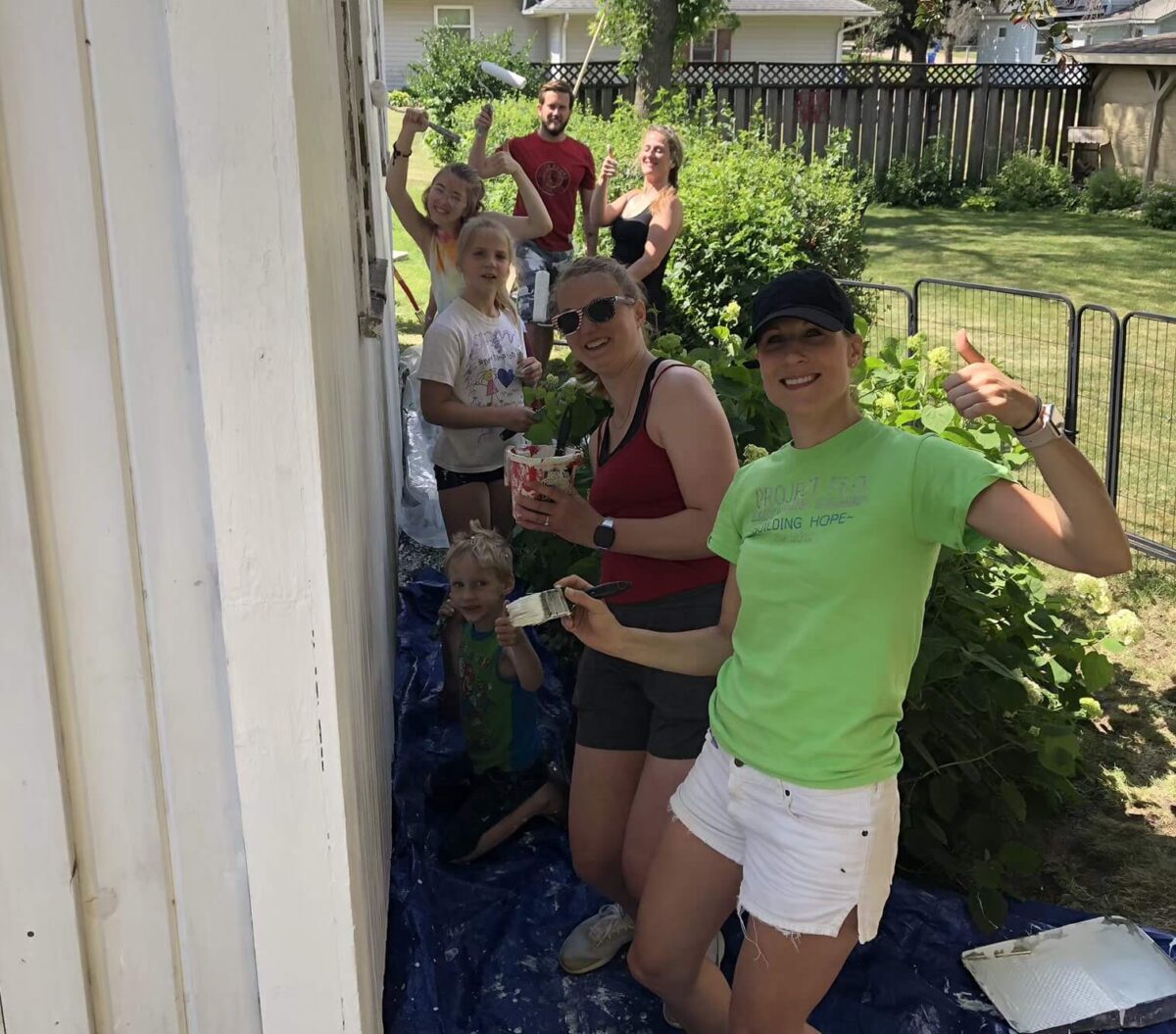 A group of people, including adults and children, paint a white wooden wall outdoors in a garden setting. One person gives a thumbs-up, and tarps cover the ground.