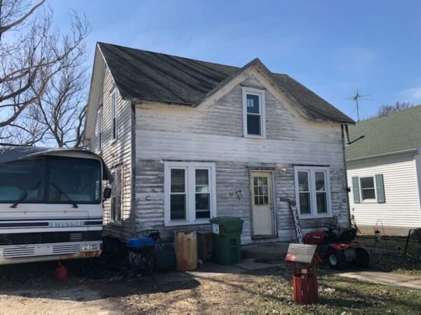 Weathered two-story house with peeling paint, adjacent to a bus and a red lawnmower, under a clear blue sky.