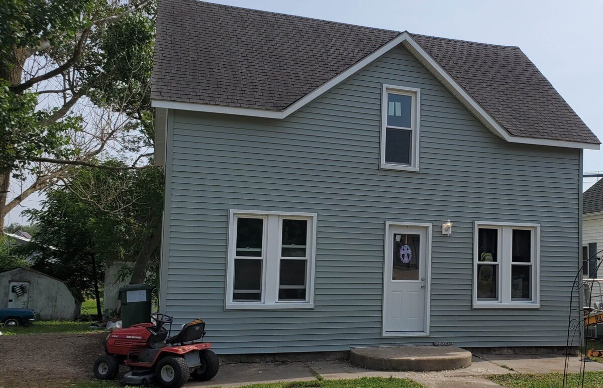 A light gray two-story house with a gable roof. A red ride-on lawnmower is parked on the concrete path leading to the white front door. Trees and a small shed are visible in the background.