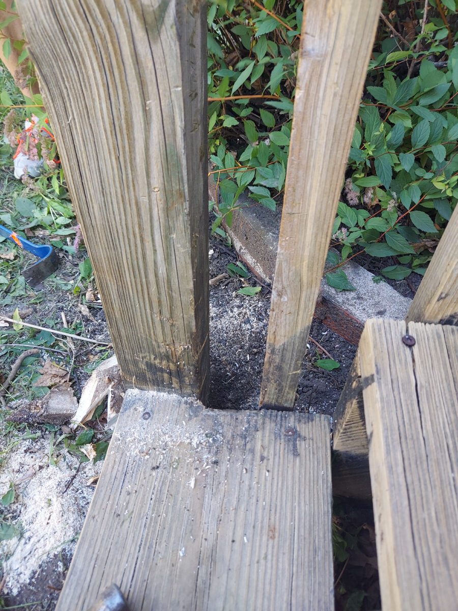 Wooden fence post with missing section, surrounded by leaves and debris, showing wood shavings and some tools in the background.