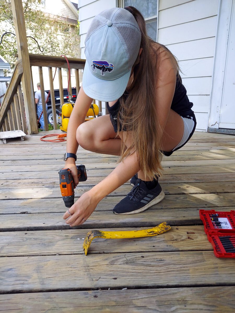 A person wearing a cap uses a power drill on wooden decking. A pry bar and a red tool case are nearby.