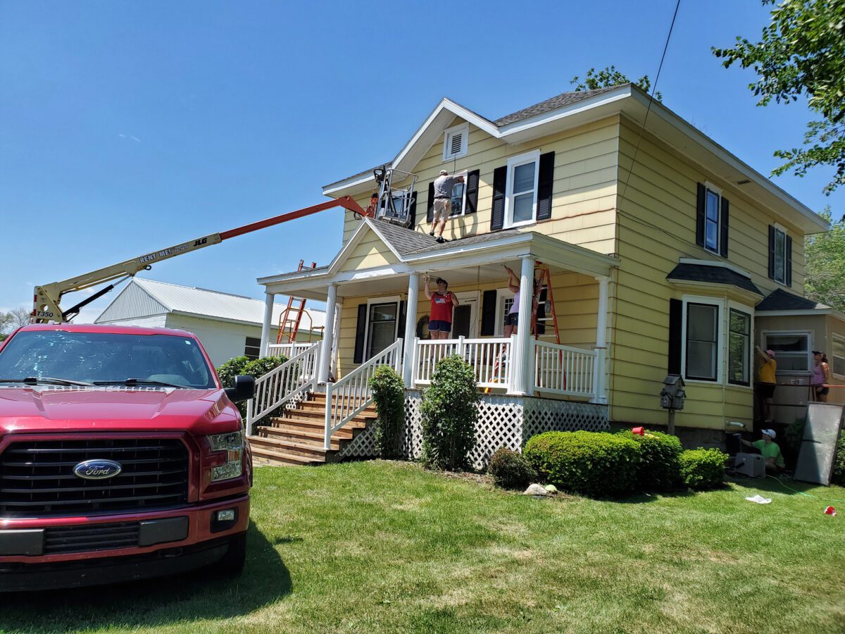 Workers on ladders and a cherry picker repair the roof and siding of a two-story yellow house with a porch. A red pickup truck is parked on the grass.