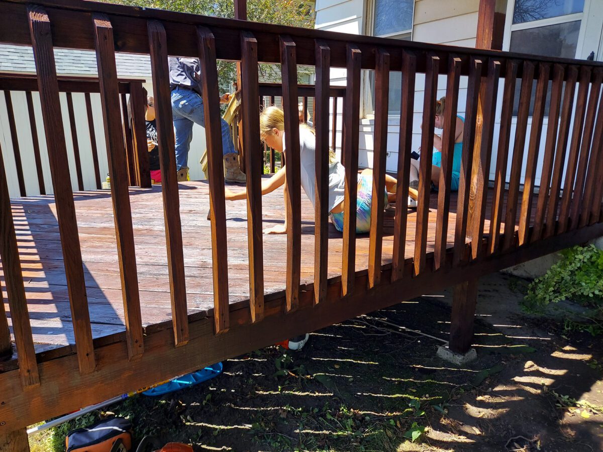 Two people staining a wooden deck with brushes under sunlight. A third person is visible partially in the background. The deck has vertical railings and is attached to a building.