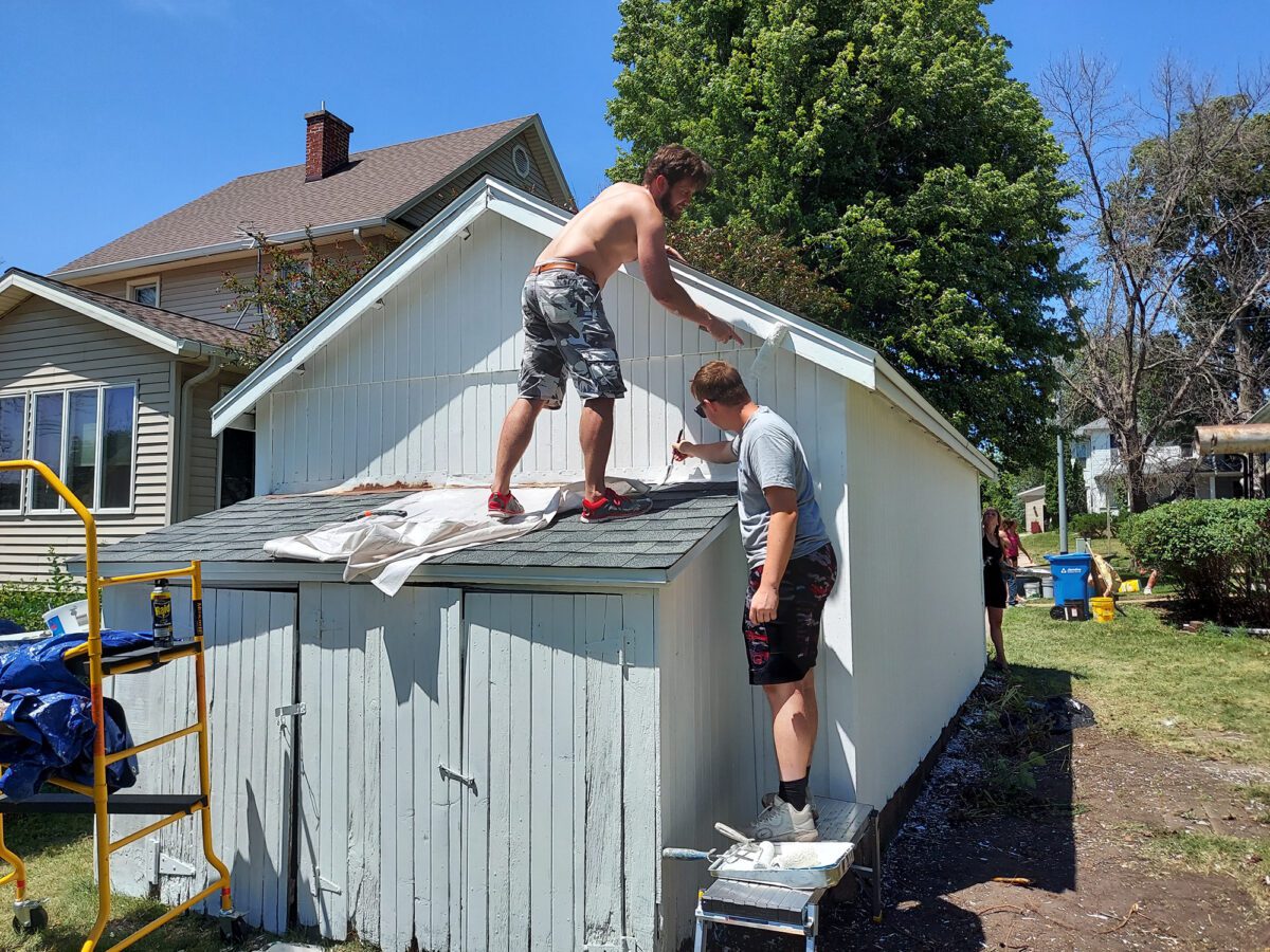 Two people painting a light-colored shed on a sunny day. One is standing on a sloped roof, while the other is on a step stool. Nearby houses and trees are visible in the background.