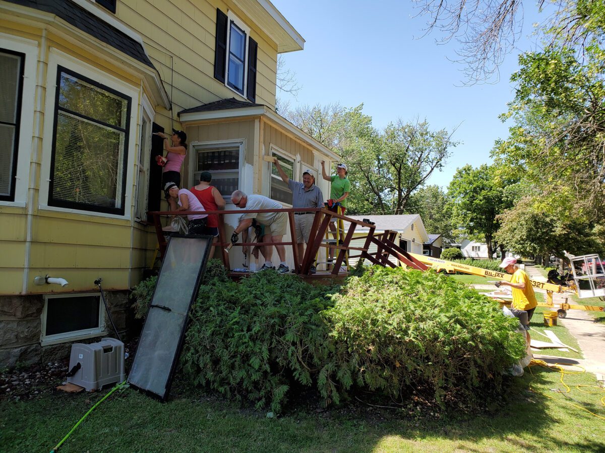 A group of people work together to remove a wooden deck from the side of a yellow house on a sunny day.