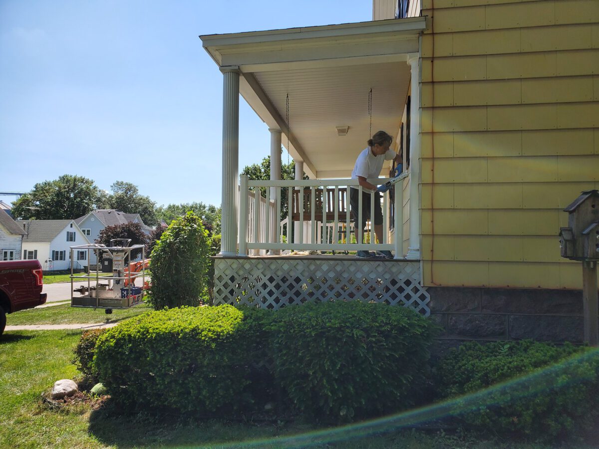 A person paints the railing of a yellow house's porch on a sunny day. Green bushes and a red vehicle are visible in the foreground.