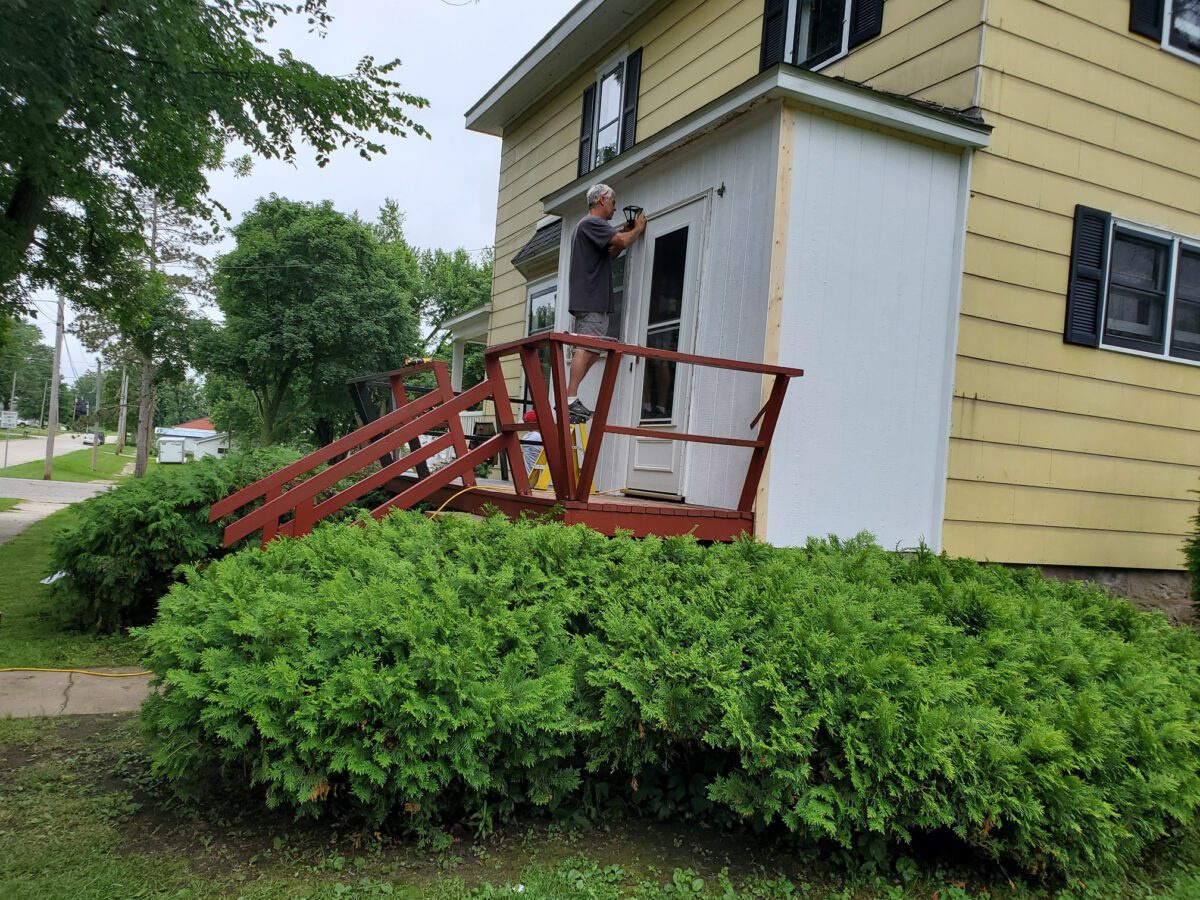 Person standing on a wooden ramp beside a yellow house, working on an exterior wall with a tool.