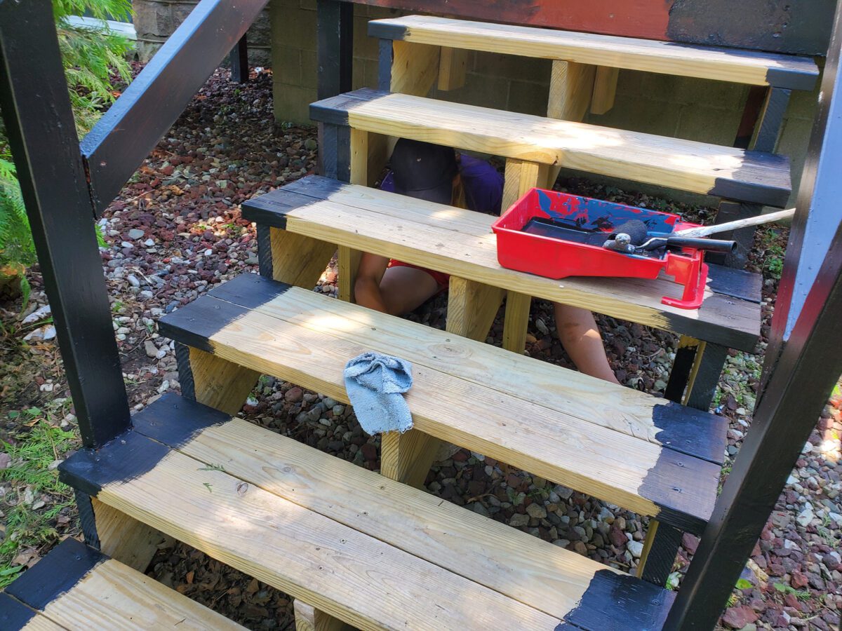 Person sitting under wooden stairs with paint tray and roller. Stairs partially painted black with a glove resting on a step.