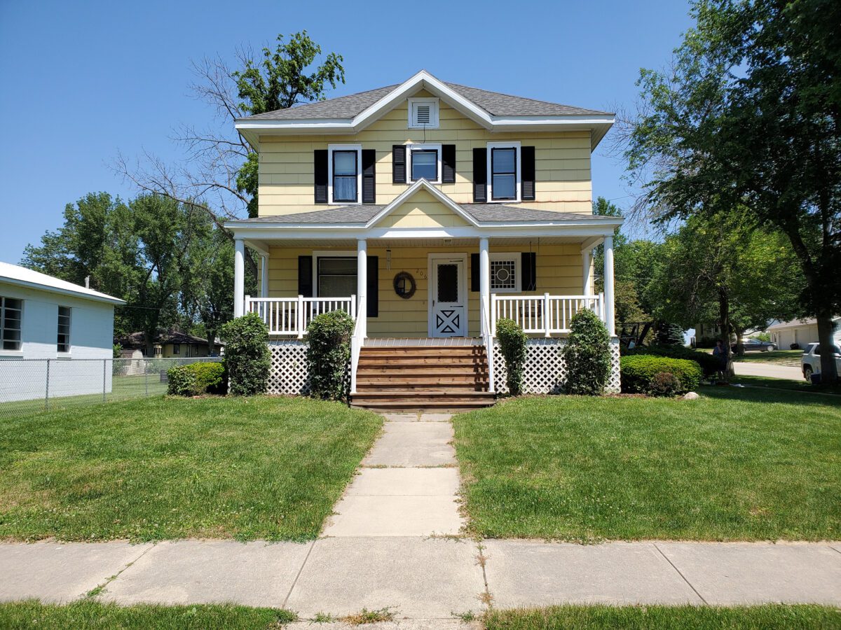 Two-story yellow house with black shutters, a covered front porch, and a white railing. The house is surrounded by a green lawn and trees on a clear, sunny day.