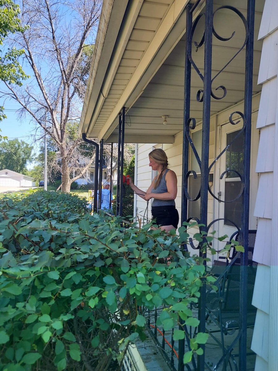 A woman stands on a porch beside a bush, painting a metal pillar. She is holding a paintbrush and wearing a tank top and shorts.