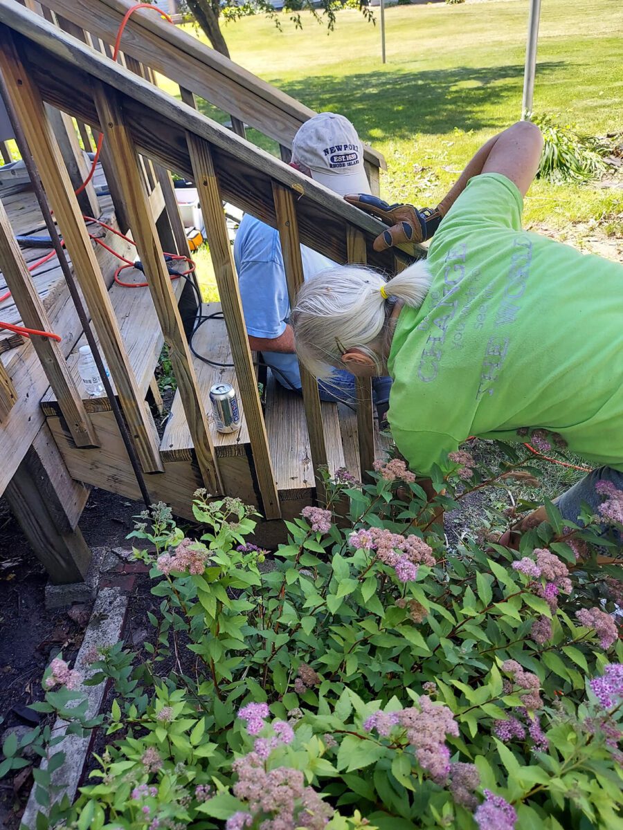 Two people repairing wooden steps outdoors, surrounded by greenery and pink flowers. A can and tools are visible nearby.