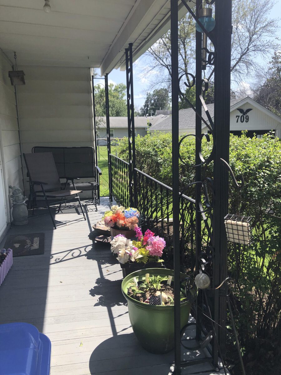 A porch with two chairs, a small table, colorful flowers in pots, and decorative metal railing. The background shows a grassy area and a house with the number 709.