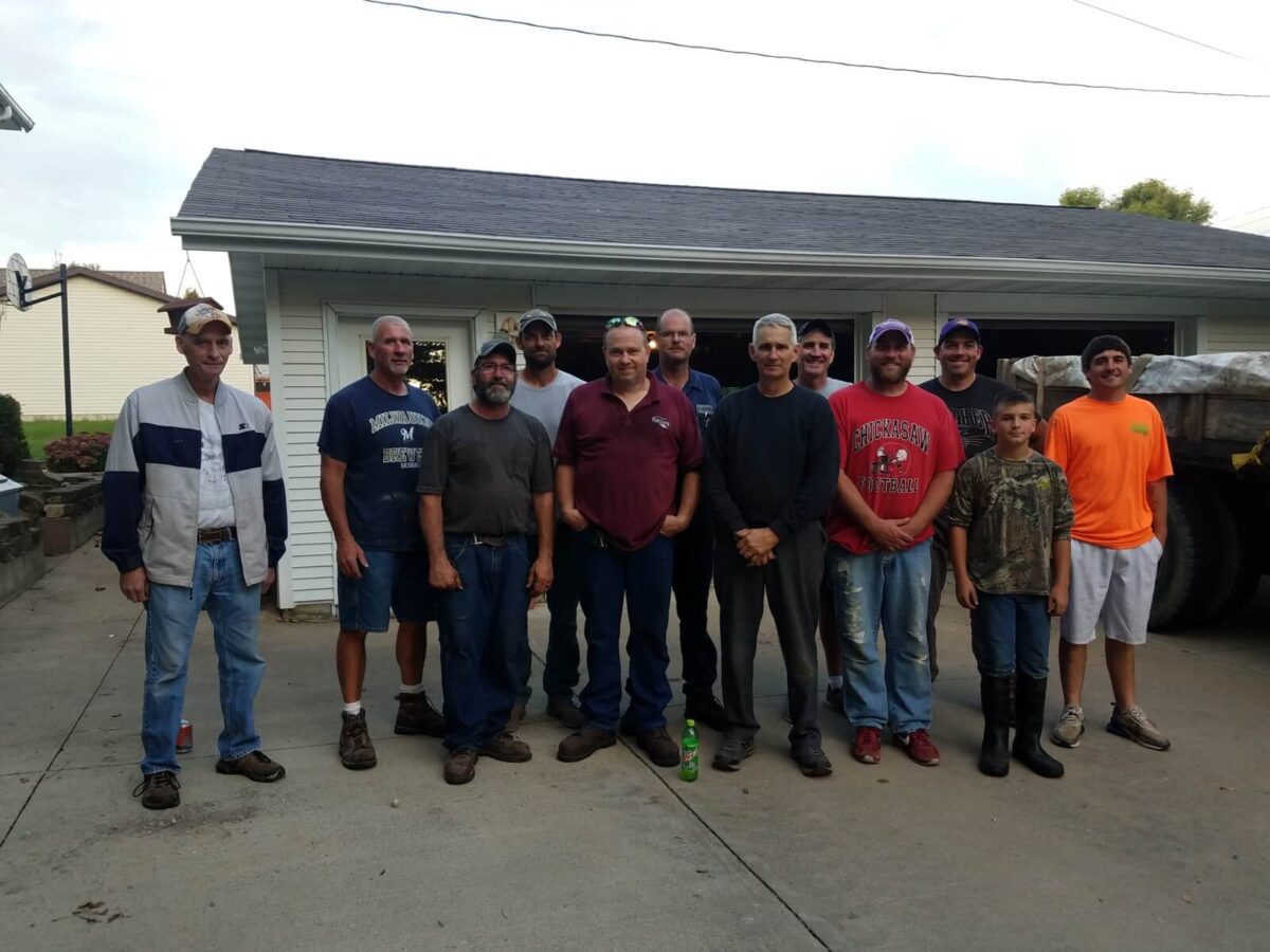 A group of thirteen people standing on a driveway in front of a garage, wearing casual clothing.