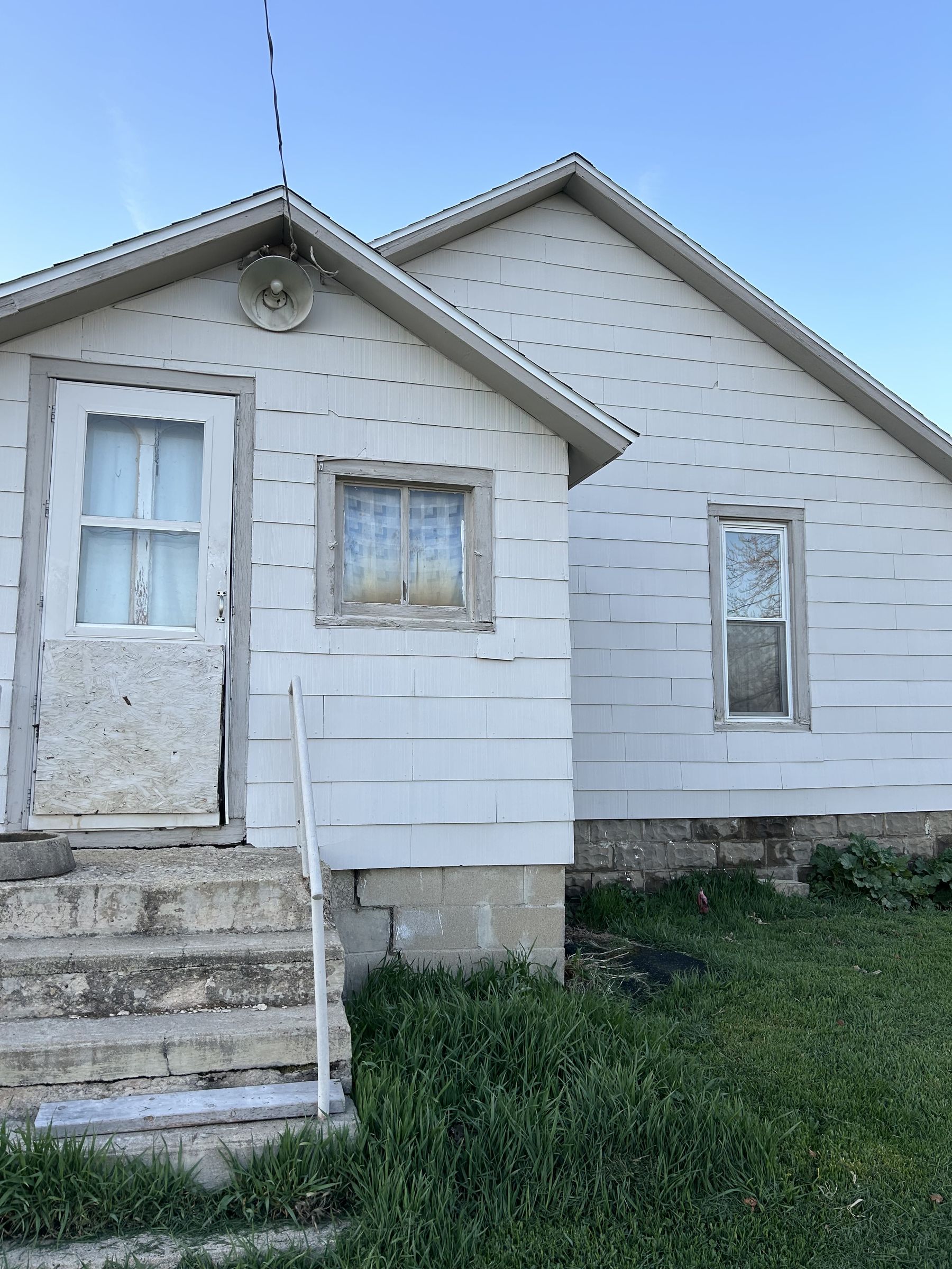 A small white house with weathered siding. A boarded-up door and two windows are visible. Steps with a metal railing lead to the entrance. Grass is growing around the foundation.