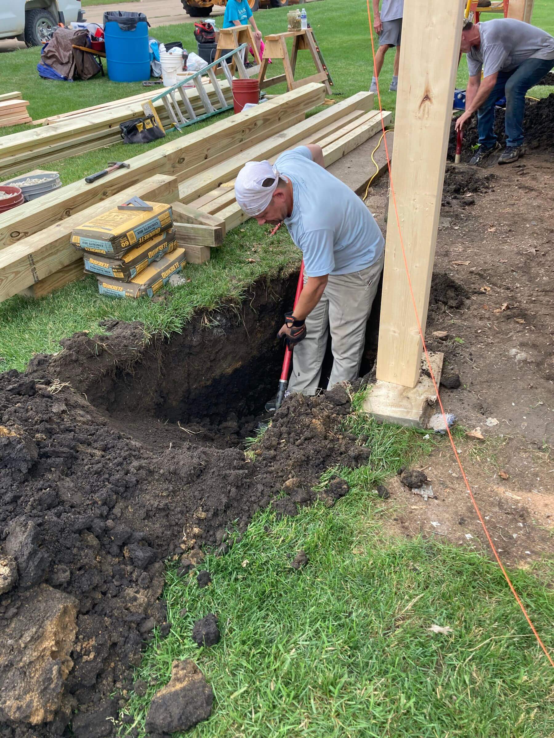 A person digs a deep hole next to a wooden post on a grassy area, surrounded by construction materials and equipment.