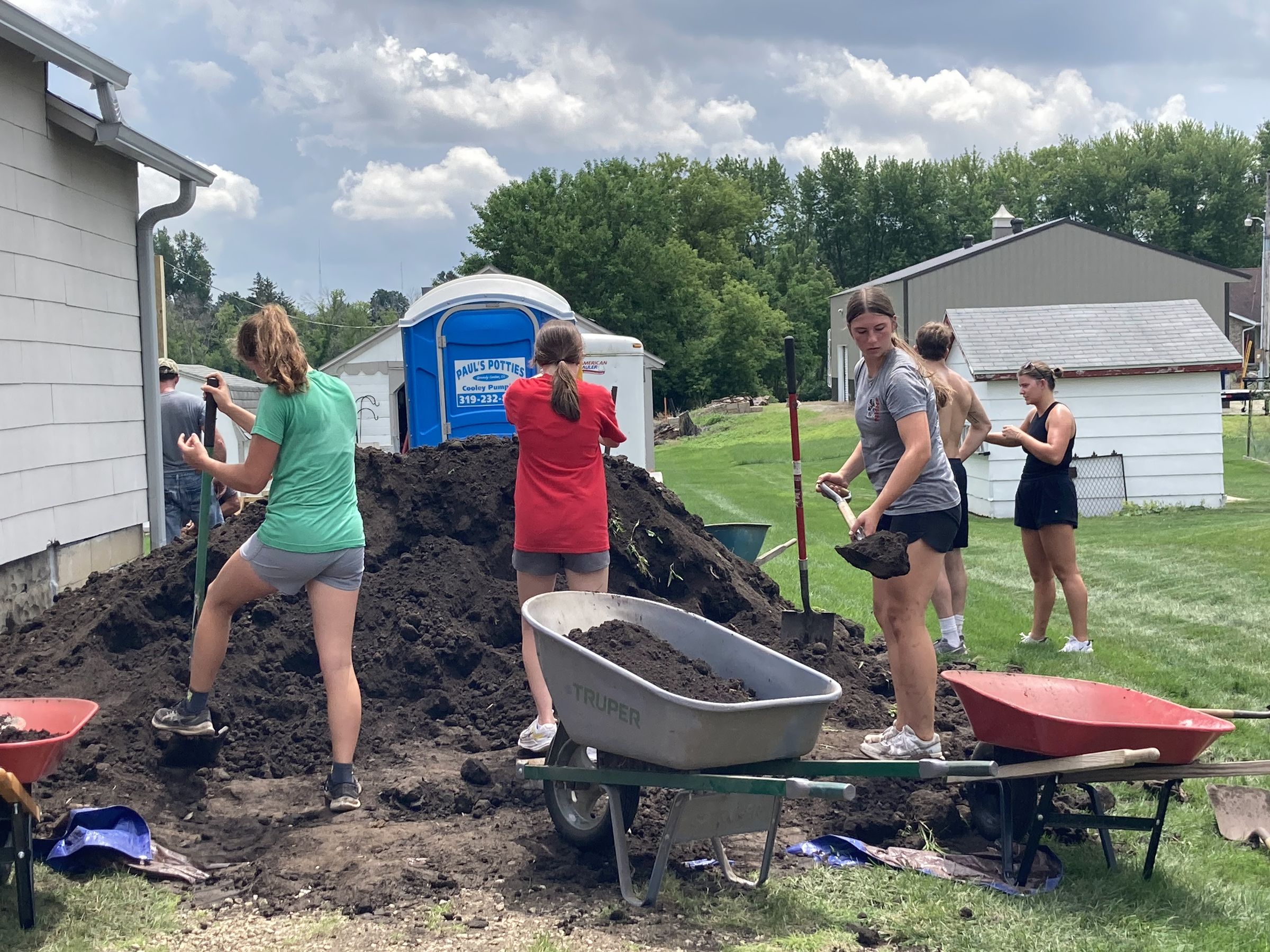 Group of people shoveling dirt from a large pile into wheelbarrows beside a building on a grassy field, with a portable toilet visible in the background.