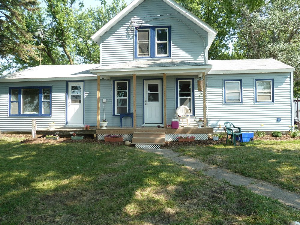 A light blue two-story house with a front porch, three visible doors, and several windows. The yard has grass and trees, with chairs placed near the porch.