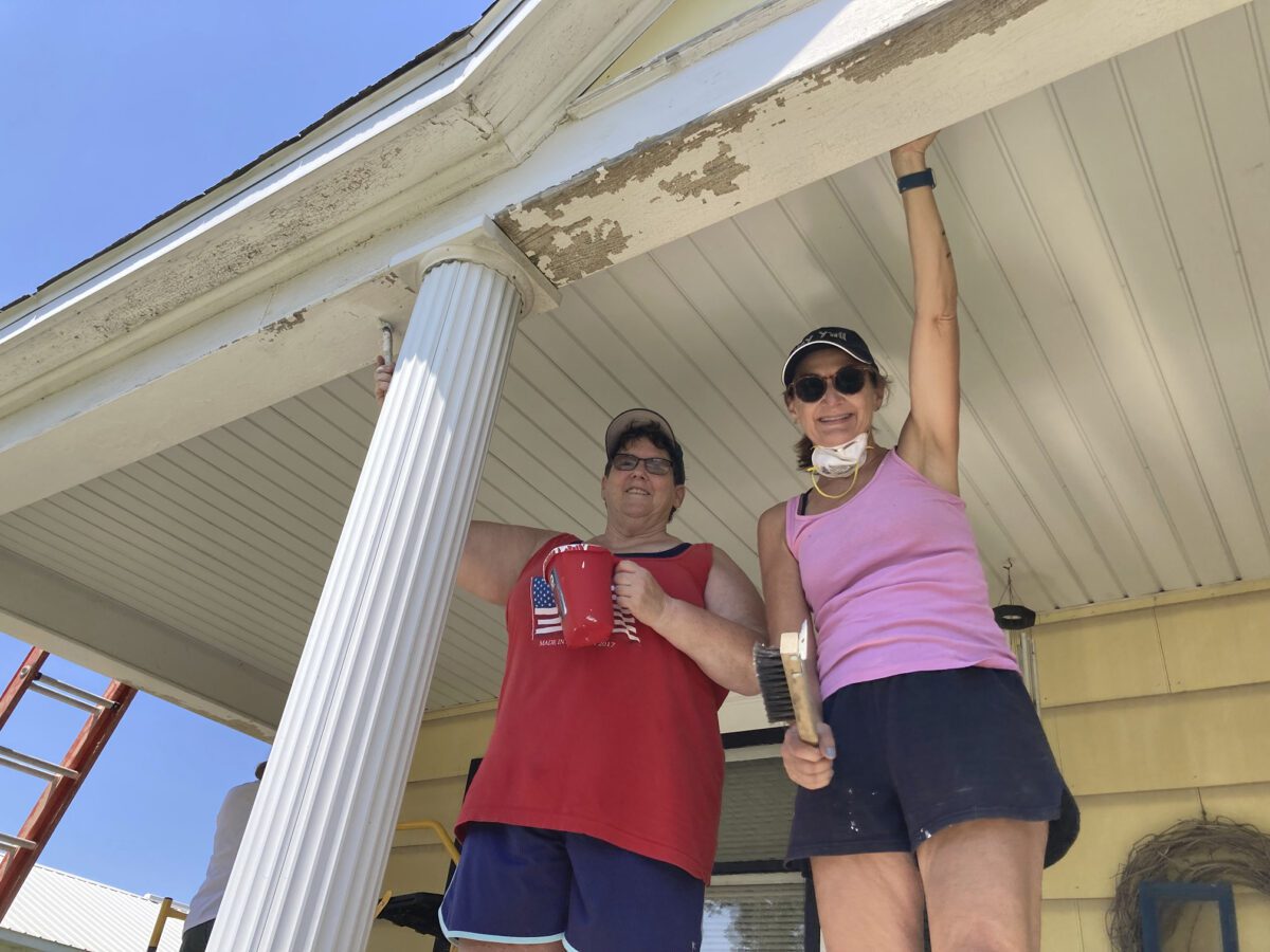 Two people are standing on a porch, holding painting tools, looking up at the eaves of a house. They appear to be involved in a home improvement project.