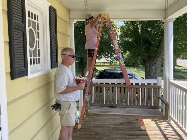 Two people work on a porch. One stands on a ladder adjusting something on the ceiling; the other holds tools, standing beside a porch swing.