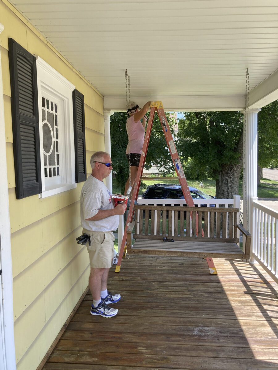 Two people work on a porch. One stands on a ladder adjusting something on the ceiling; the other holds tools, standing beside a porch swing.