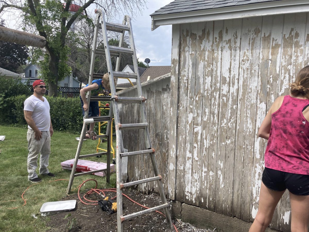 People working on painting a weathered wooden building, with ladders and paint supplies scattered around.