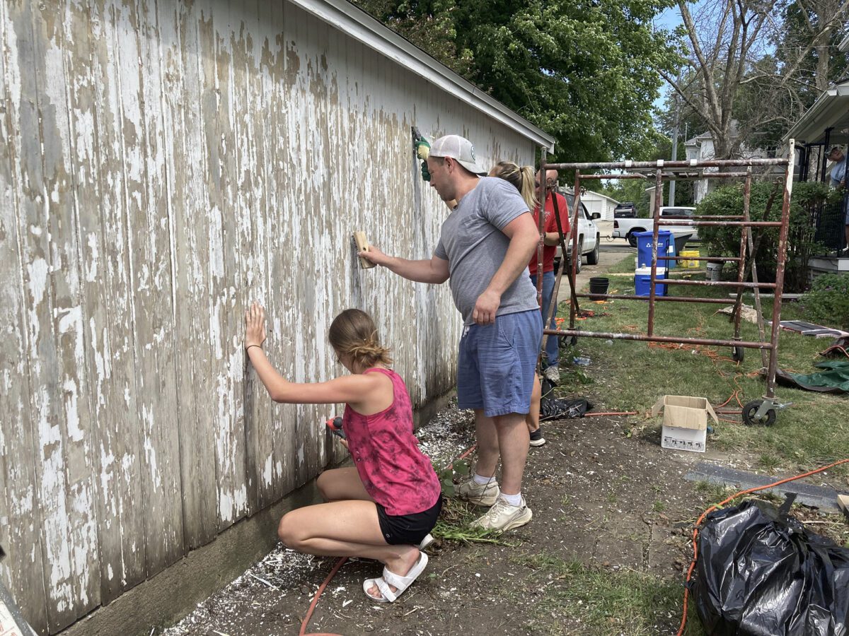 People removing old paint from a wall outdoors. A man and a woman work with tools on the worn surface. Scaffolding and materials are visible nearby.