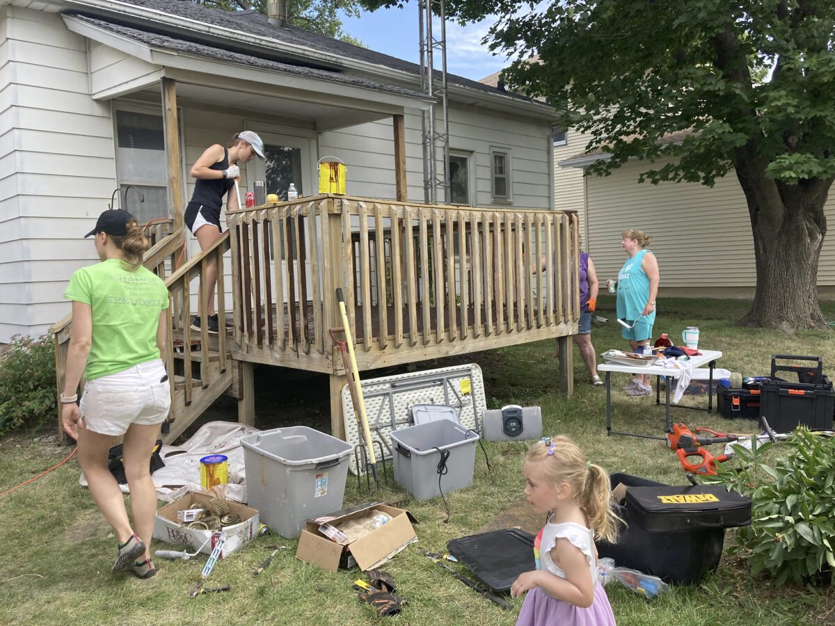 People working on a wooden porch, surrounded by tools and supplies. A woman in a green shirt walks away while a child stands in the foreground.