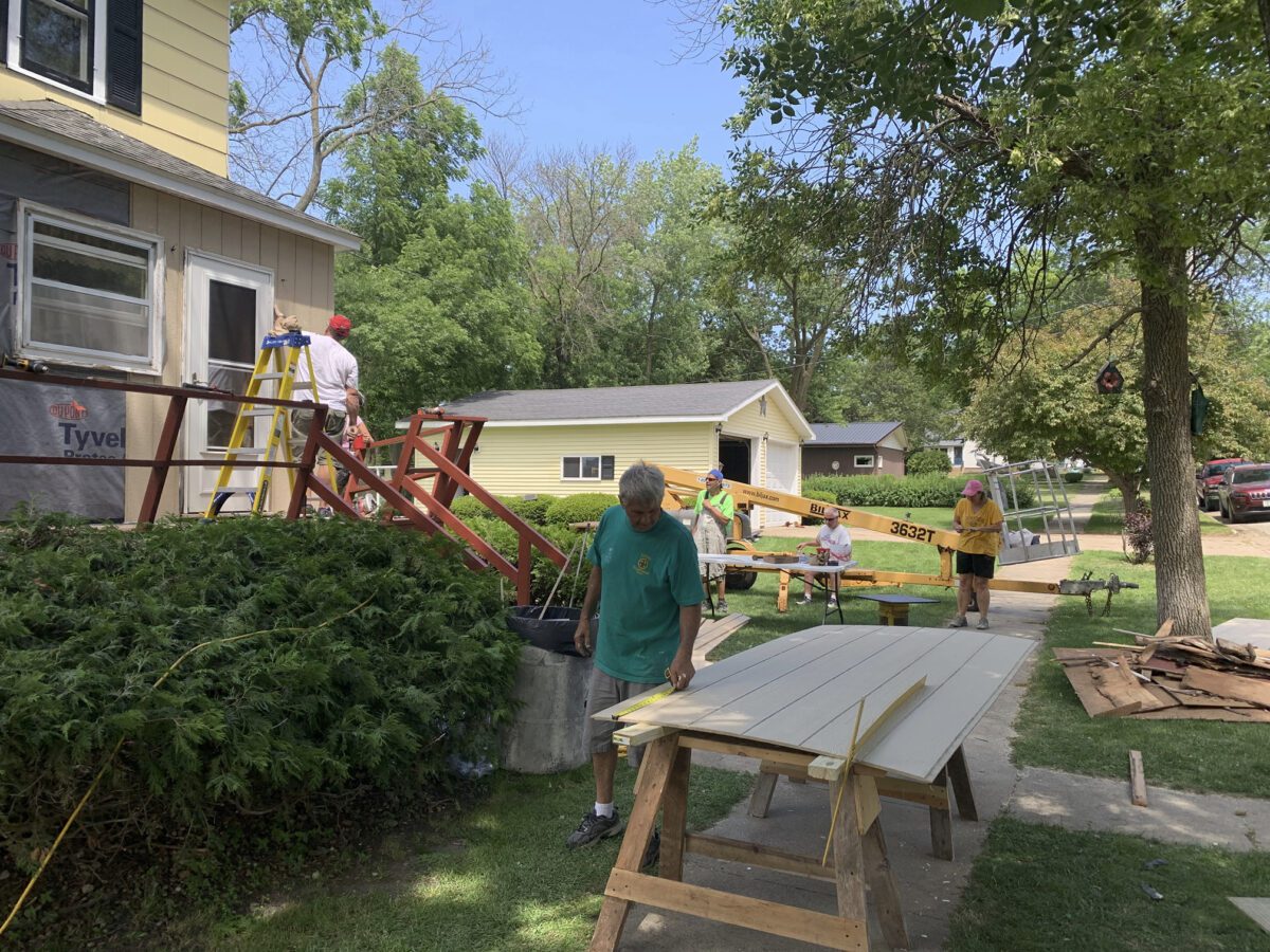 People working on home repairs outside a house. One is on a ladder by the house while others handle materials on the ground. Trees and a neighboring house are in the background.