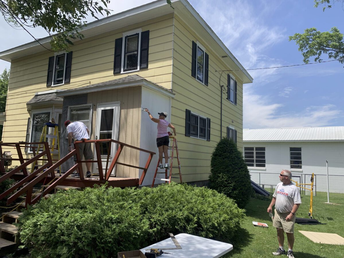 Three people work on painting the exterior of a yellow two-story house. Two are on ladders painting, while another person stands nearby. A table and some tools are visible on the grass.