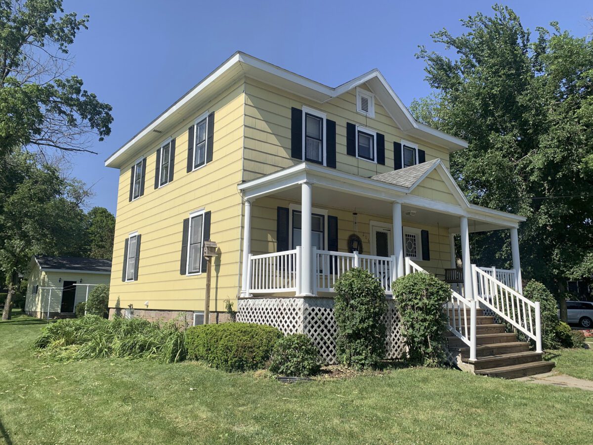 Yellow two-story house with a white front porch and black shutters, surrounded by green lawn and bushes, under a clear blue sky.