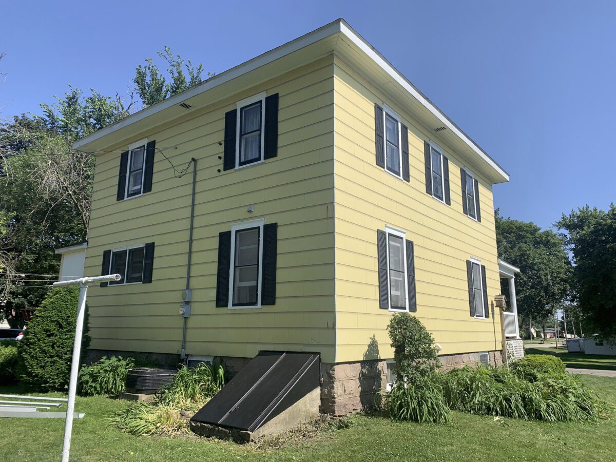 A two-story yellow house with black shutters, surrounded by greenery, under a clear blue sky. Steps lead to a basement entrance on the side.