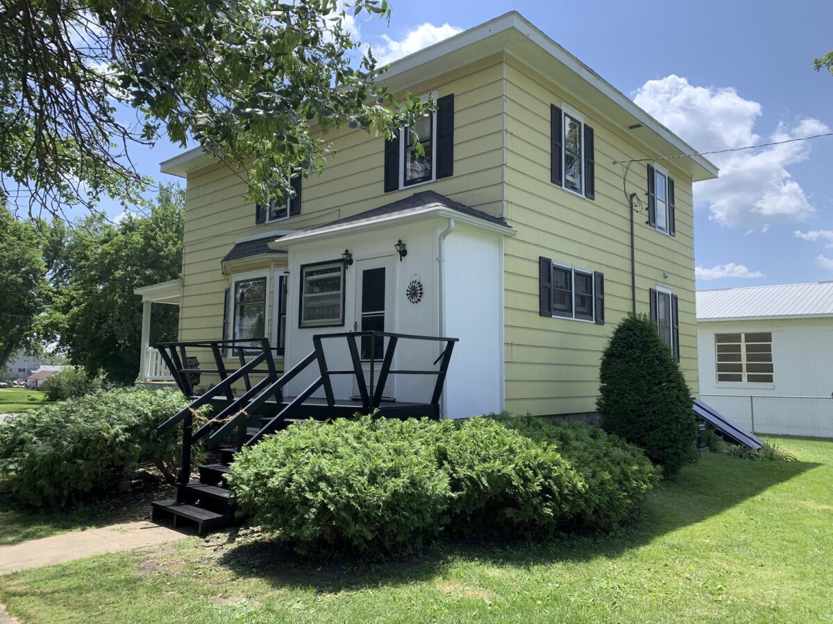 Two-story yellow house with a black porch railing, white entrance, and well-kept bushes, set in a grassy yard under clear blue skies.