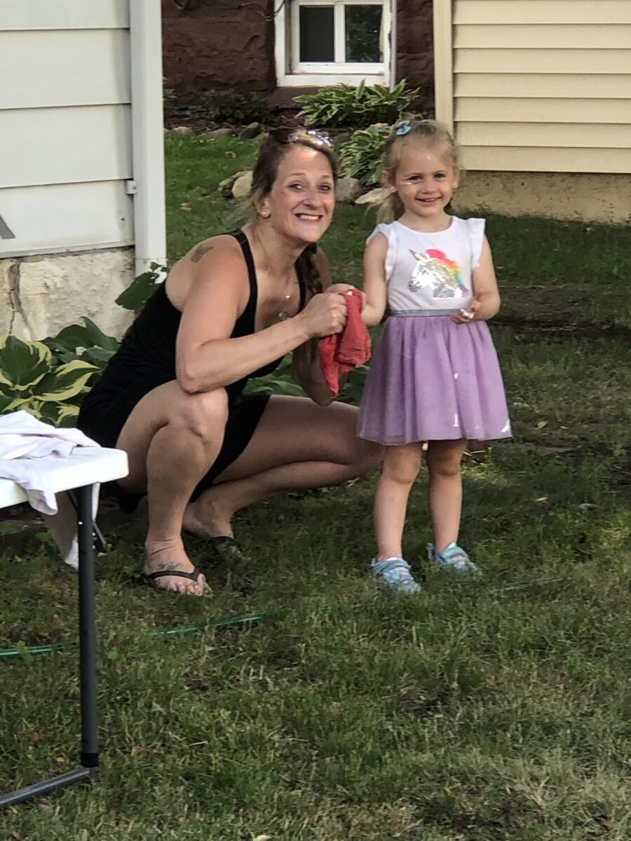 A woman squats next to a smiling young girl in a purple skirt on a grassy area near a building.