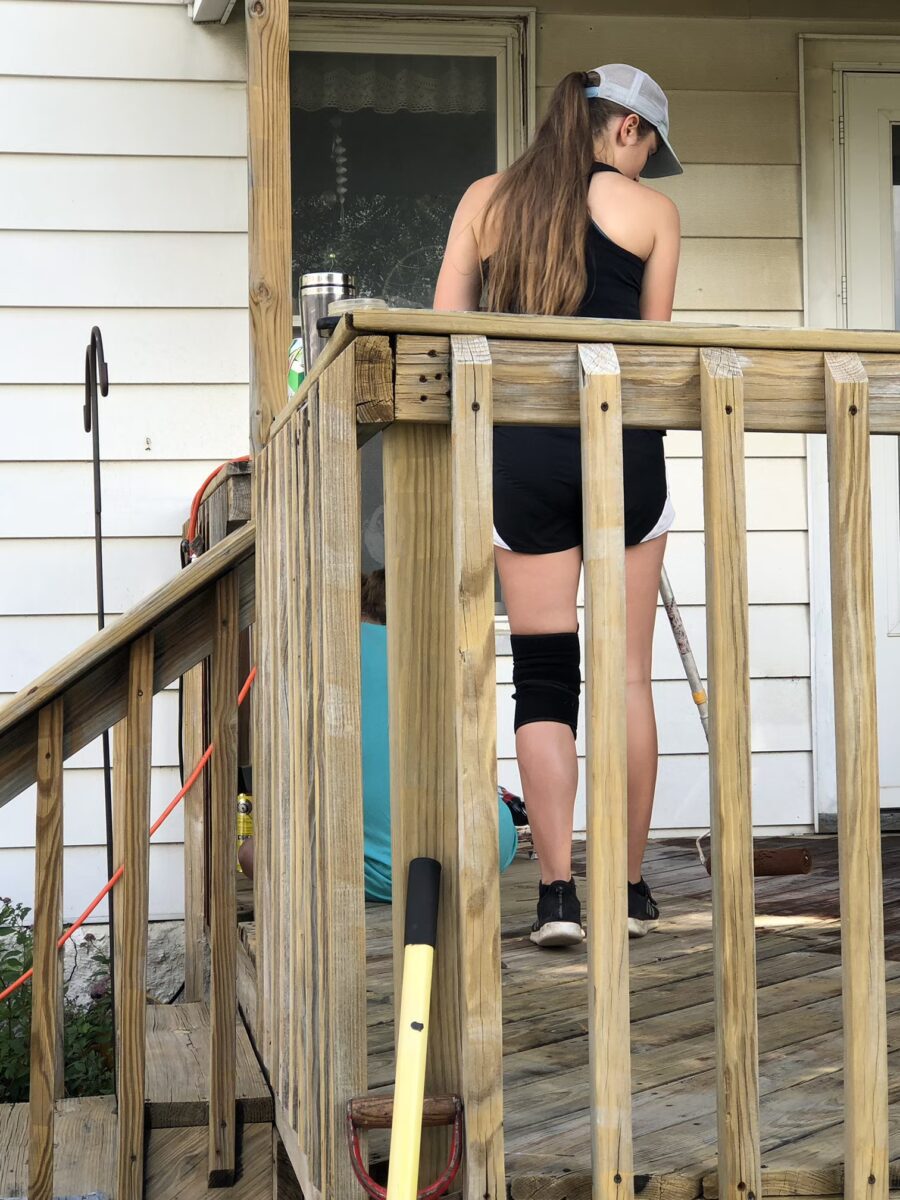 Person wearing a cap, tank top, and knee brace, standing on a wooden porch with cleaning tools.