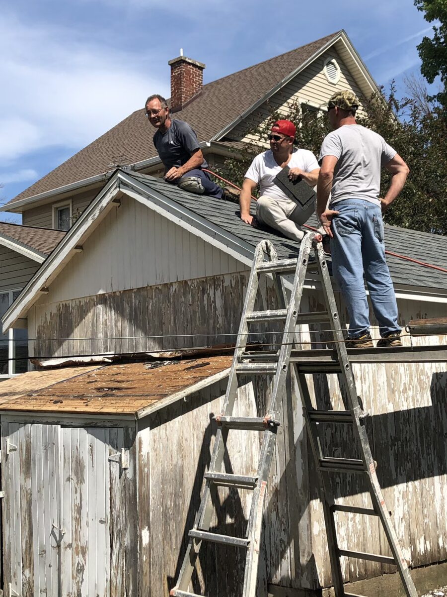 Three men work on the roof of a weathered garage, with ladders positioned for access. A house is visible in the background under a clear blue sky.