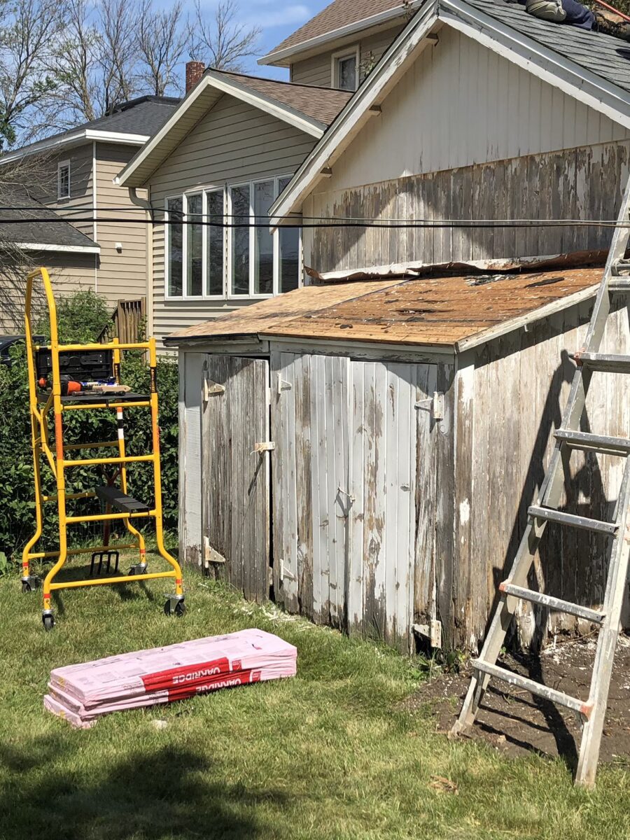 A weathered wooden shed with peeling paint and missing roof panels. Ladders and a yellow scaffold are positioned nearby. Pink roof shingles lie on the grass.