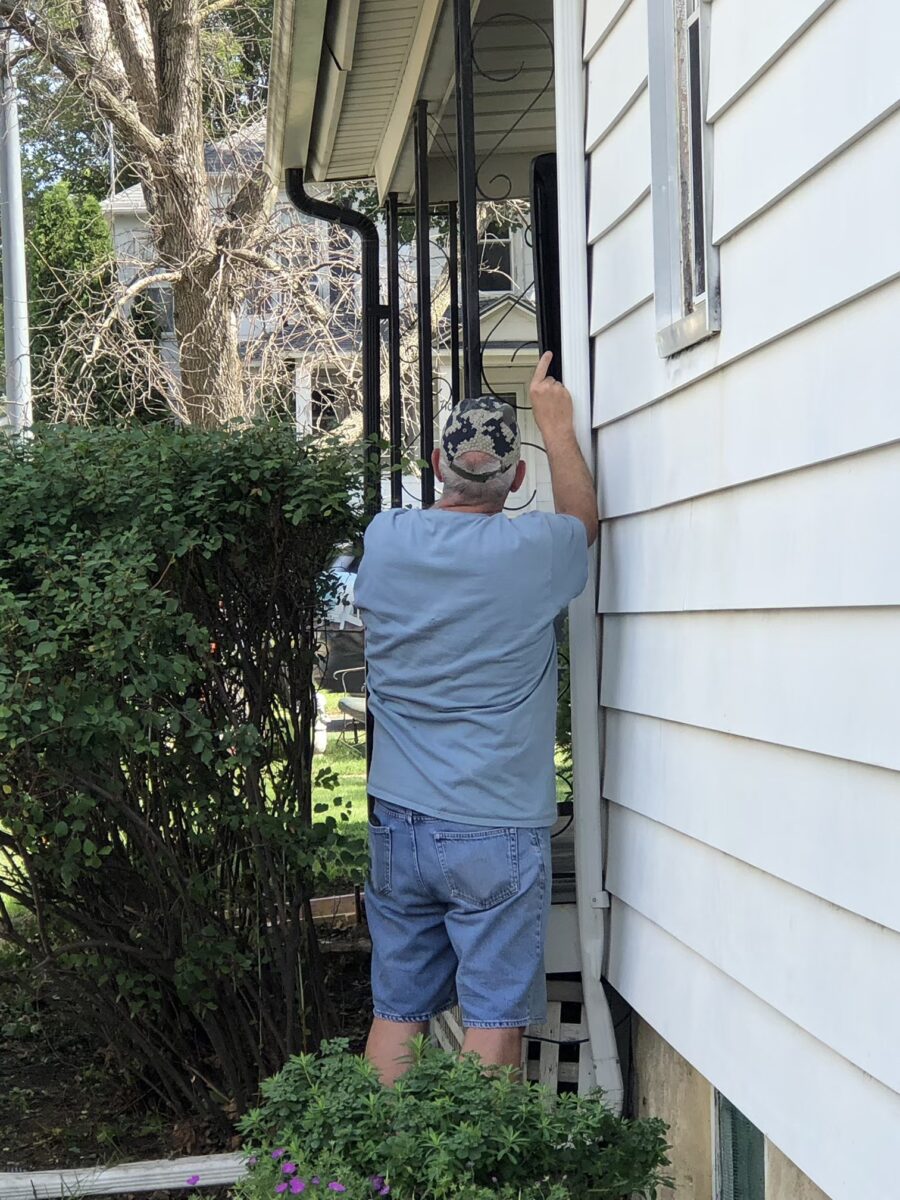 Person in a camouflage cap and blue shirt standing outside a house, reaching towards a window frame. Lush greenery and a white siding are visible.