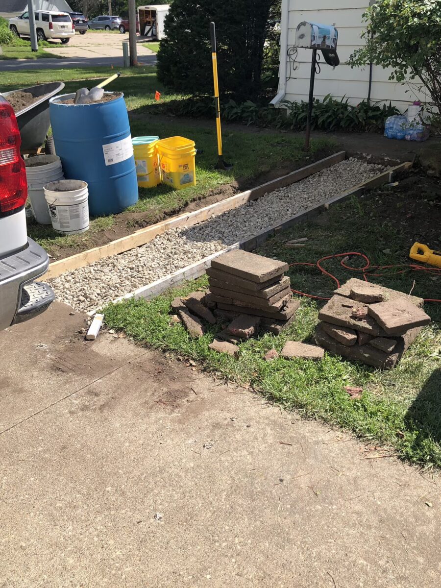 Pavers stacked on grass beside a gravel path under construction next to a driveway with tools and buckets nearby.