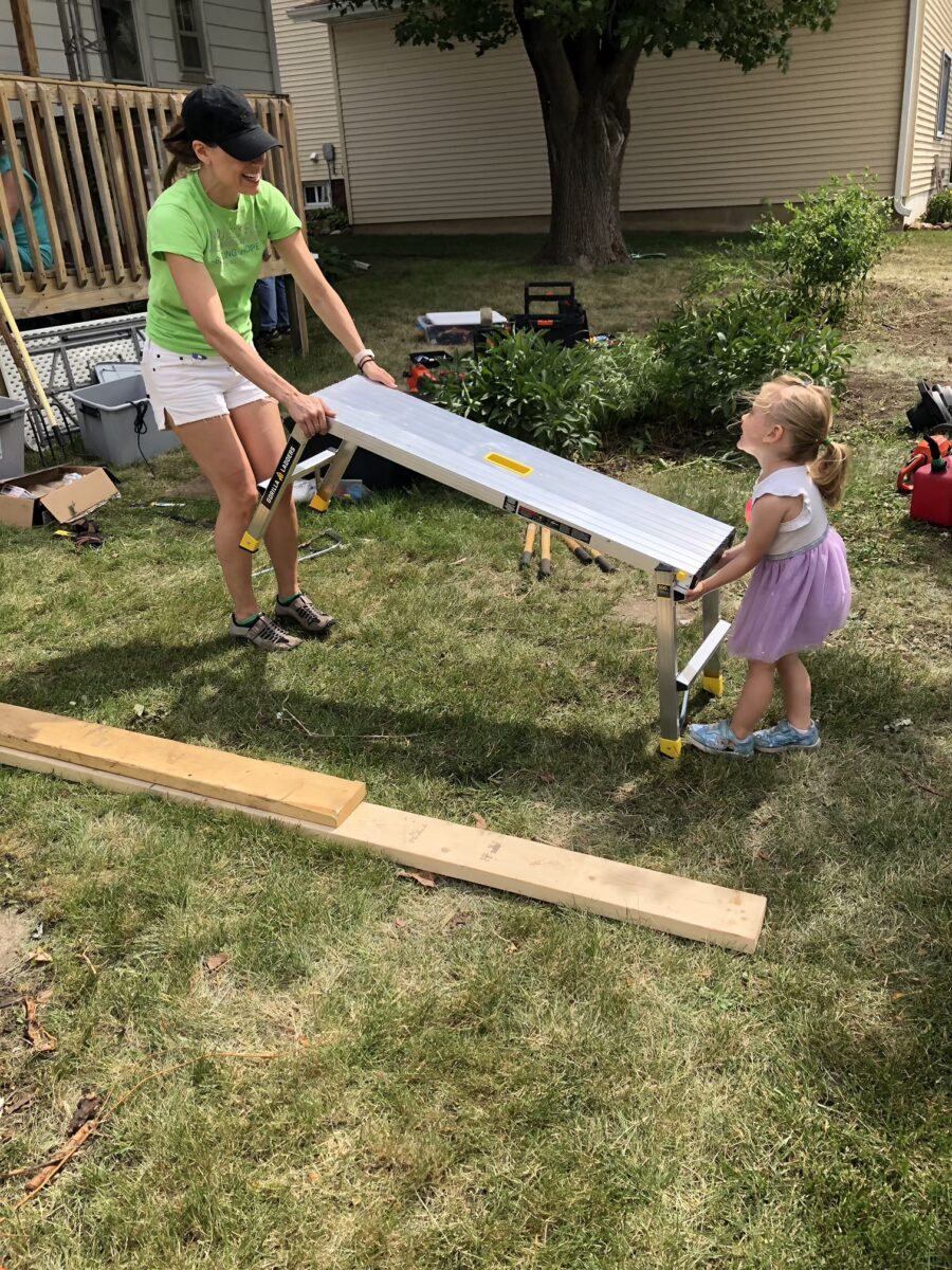 An adult and a child hold opposite ends of a folding ladder outdoors on grass, surrounded by tools and wooden planks.