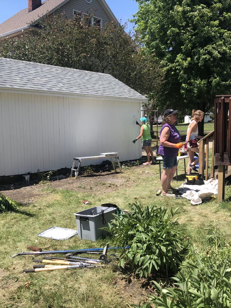 People painting the side of a white garage, surrounded by grass and garden tools, on a sunny day.