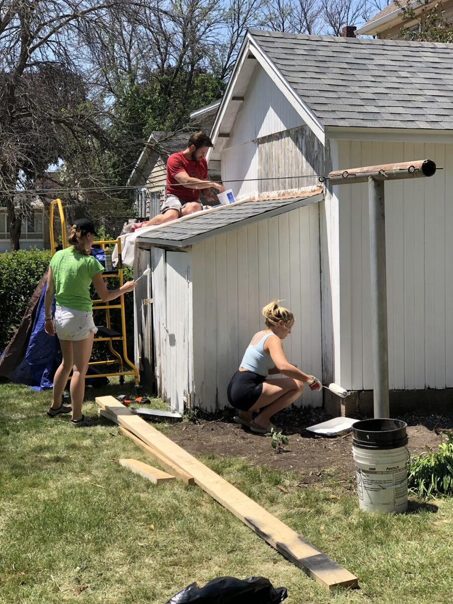 Three people repaint a small white building. One is on the roof, another on a scaffold, and the third is painting near the base. Wooden planks and supplies are on the grass.