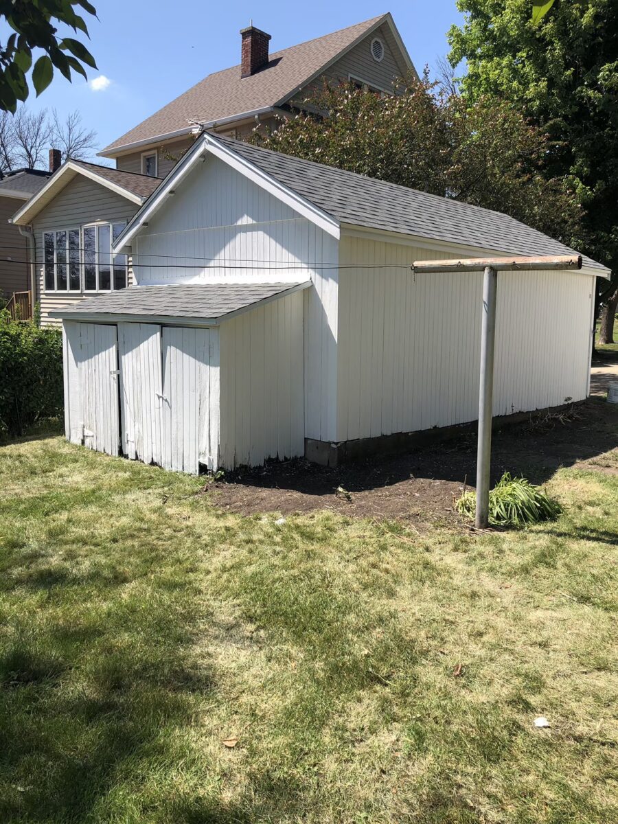 A white wooden shed with three doors stands on a grassy backyard. A bare clothesline pole is visible in the foreground, and a house with large windows is in the background.