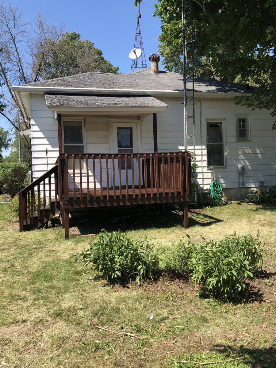 A small white house with a gray roof and a wooden deck. A garden hose is coiled on the wall, and a patch of greenery is in the foreground. Trees and a windmill are visible.