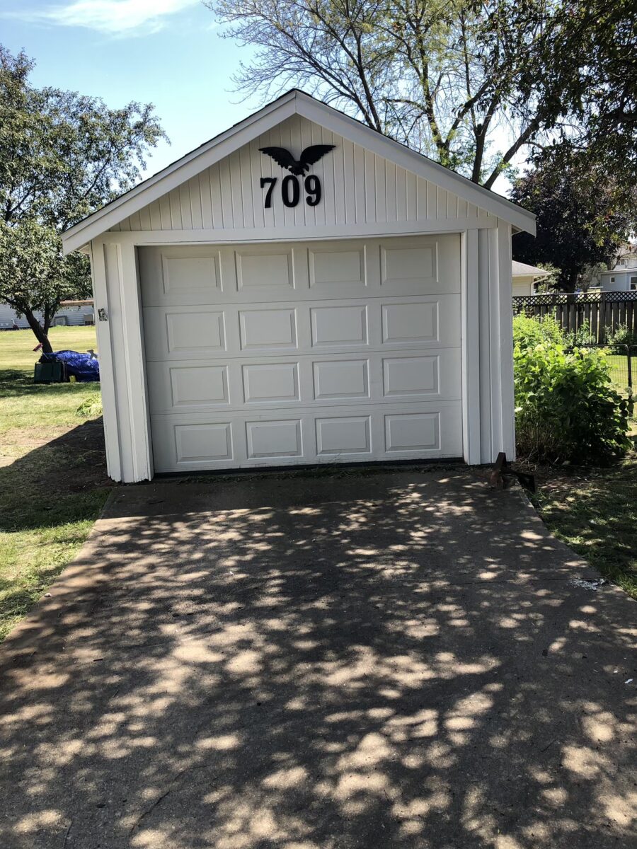 A white garage with the number 709 and a decorative black butterfly above the door. There's a driveway in front, and trees cast shadows on the ground.